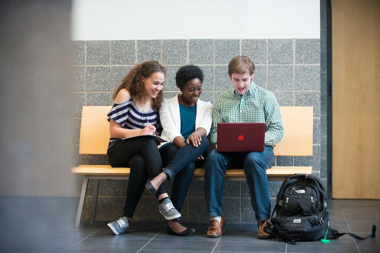 Three students sitting on bench looking at red laptop