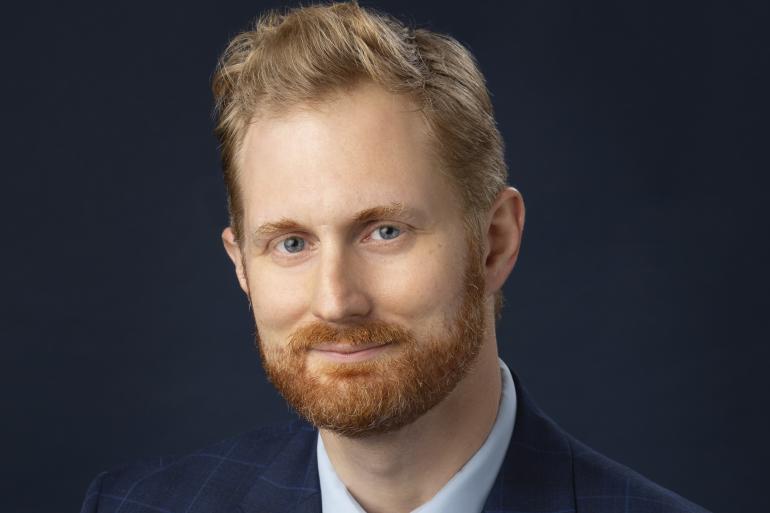 Jason Turkiela is pictured from the sternum up. He has blonde-ginger hair and beard, and is wearing a dark blue suit with a red and blue striped tie.