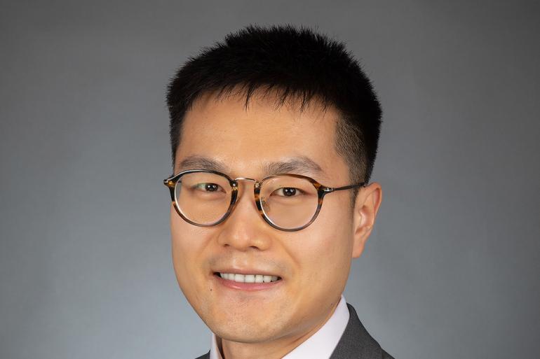 Adam Zhang is picture from the sternum up. He has black hair and glasses. He is wearing a dark gray suit with a black tie.
